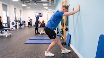 Hombre en ropa deportiva estira contra pared. En el fondo, otros individuos y equipos de gimnasio. Dos colchonetas y bandas elásticas en la sala.