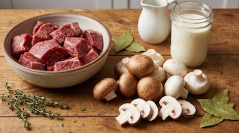 Un tazón con carne de res en cubos, champiñones enteros y rebanados, un frasco de crema de leche, laurel y tomillo sobre una superficie de madera.