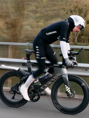 Ciclista vestido de negro con casco blanco pedaleando una bicicleta de carretera oscura con ruedas de disco y guardabarros. Fondo borroso con árboles