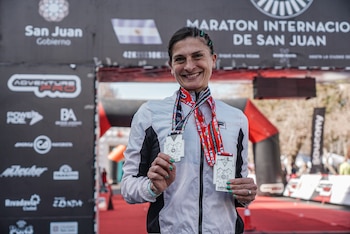 Primer plano de una mujer sonriente con cabello trenzado y uñas verdes que sostiene dos medallas de plata. De fondo hay un banner oscuro con texto y la bandera argentina