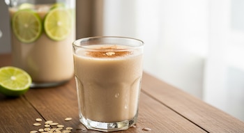 Vaso de agua de avena cremosa con canela en polvo sobre una mesa de madera. Un limón partido y hojuelas de avena esparcidas. Jarra al fondo.
