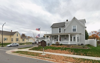 Calle residencial con casa gris de dos pisos y porche blanco, mástiles con bandera de Estados Unidos y de Ohio, placa histórica, jardín con hojas secas