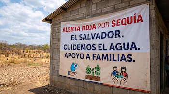 Lona informativa colgada en una pared de bloques de hormigón junto a un terreno de tierra seca y agrietada.