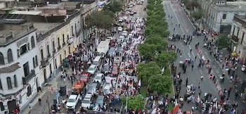 Vista aérea de una calle en ciudad. Edificios de época a los lados. Calzada ocupada por una marcha de personas, algunas con banderas. Vehículos estacionados y en circulación. Bulevar con árboles