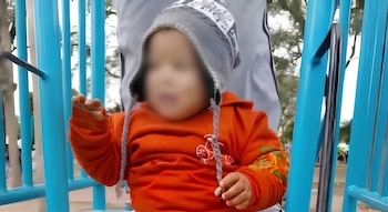 Un niño pequeño con la cara difuminada viste una sudadera naranja y gorro gris entre estructuras metálicas azules