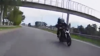 Vista desde el frente de un motociclista con casco y chaqueta oscura conduciendo por una autopista gris, bajo un puente peatonal elevado y con árboles a los lados