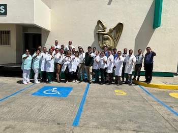 Un grupo de profesionales de la salud del IMSS, con batas y uniformes, posa con pulgares arriba frente a un edificio con el logo institucional y un espacio para discapacitados