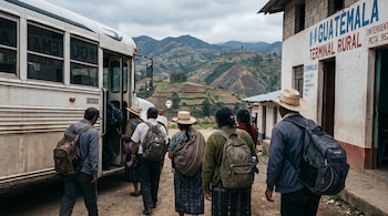 Grupo de hombres y mujeres, algunos con sombreros y vestimenta tradicional, desciende de un autobús blanco frente a una terminal rural en Guatemala, con montañas y cielo nublado.
