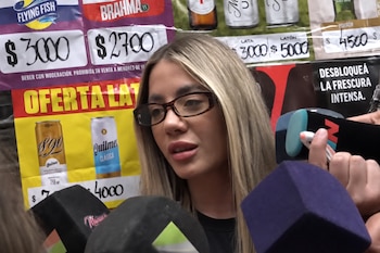 Una mujer rubia con lentes habla frente a micrófonos de prensa ante un fondo con anuncios de bebidas y precios.