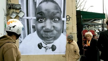 Un gran retrato en blanco y negro de un rostro con ojos grandes y labios fruncidos pegado en una pared, con peatones y cajas eléctricas visibles