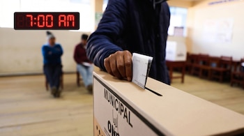 Primer plano de una persona depositando una papeleta en una urna de elecciones municipales. Un reloj digital en la esquina superior izquierda marca las 7:00 AM.