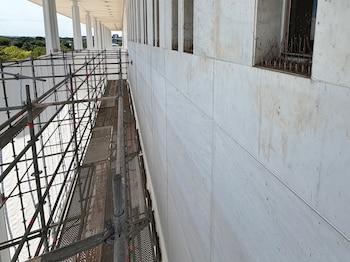 Andamios metálicos junto a la fachada de concreto de un edificio. Múltiples aberturas con rejillas de metal. Cielo azul y árboles en la distancia