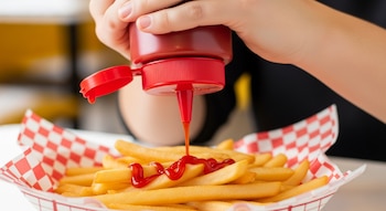 Primer plano de una mano exprimiendo una botella roja de kétchup sobre una cesta llena de papas fritas doradas con un forro de papel a cuadros rojos y blancos.