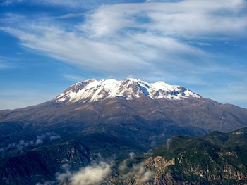 Una gran montaña con cumbre nevada y laderas rocosas bajo un cielo azul con nubes delgadas
