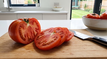 Un tomate cortado a la mitad y en rodajas sobre una tabla de madera con un cuchillo en un mostrador de cocina.