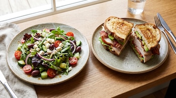 Dos platos sobre una mesa de madera, uno con ensalada verde, tomates, pepino, aceitunas y queso, y otro con un sándwich de pavo, tocino y aguacate.