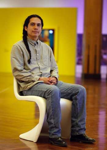 Un hombre joven de cabello largo trenzado, viste camisa clara y jeans, sentado en una silla blanca moderna. Fondo de pared amarilla y piso de madera brillante