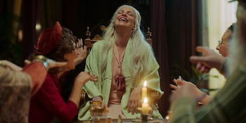 Una mujer de cabello blanco/rubio muy claro ríe alegremente con la cabeza hacia atrás, vestida con una bata verde. Está sentada en una mesa con velas y personas