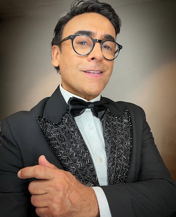 Adal Ramones sonríe a cámara, vistiendo un esmoquin negro con solapa y frente de camisa de cuentas negras, corbatín negro y gafas redondas