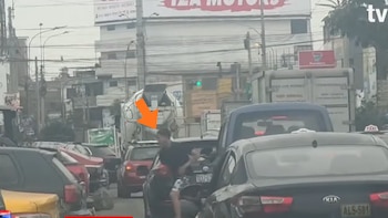 Un joven con camiseta negra y shorts es señalado por una flecha naranja en medio del tráfico vehicular en una avenida urbana, con edificios al fondo