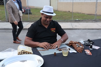 Un hombre con sombrero blanco manipula hojas de tabaco sobre una mesa con herramientas de corte, un encendedor y cigarros