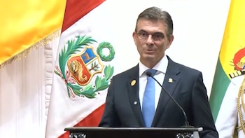 Un hombre de traje oscuro, corbata azul y gafas habla en un podio con micrófono, flanqueado por la bandera de Perú y una bandera amarilla y verde