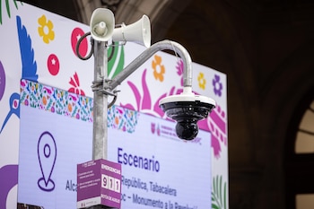 Poste metálico con dos altavoces blancos, una cámara de seguridad negra y un botón de auxilio 911 púrpura, frente a una pantalla con gráficos coloridos