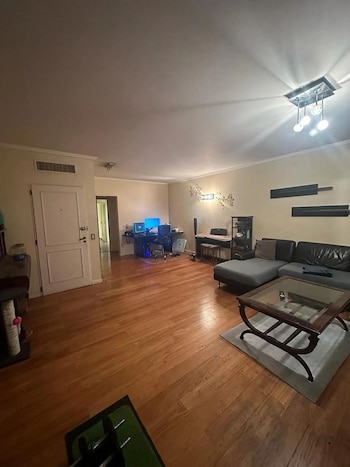 Una sala de estar con piso de madera, un sofá esquinero gris oscuro, una mesa de centro con superficie de cristal y un escritorio con dos monitores