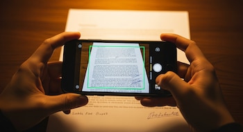 Manos sosteniendo un smartphone que muestra en su pantalla un documento físico siendo escaneado, con un recuadro verde que delinea el texto.