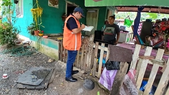 Un hombre con chaleco naranja y gorra azul escribe en un cuaderno, frente a una mujer que lo mira. Detrás, una casa turquesa, plantas y una manguera verde