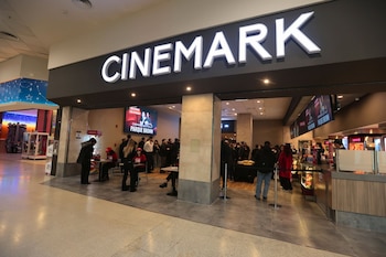 Letrero Cinemark iluminado sobre la entrada de un cine. Pantalla electrónica con información de películas. Mostrador de golosinas. Grupo de personas