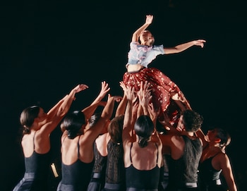 Una bailarina con blusa clara y falda roja es elevada por ocho bailarines con trajes oscuros en un escenario a oscuras, con brazos alzados hacia ella