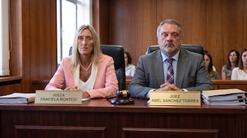 Una mujer y un hombre en traje están sentados tras un escritorio con placas identificatorias de juez, papeles y un mazo de madera.