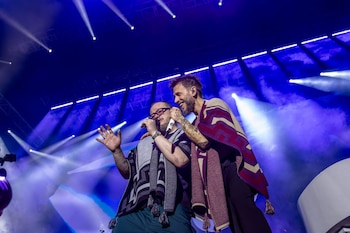 Dos hombres en un escenario cantan en micrófonos. El fondo es azul con luces y humo. Uno viste un poncho gris y otro un poncho rojizo