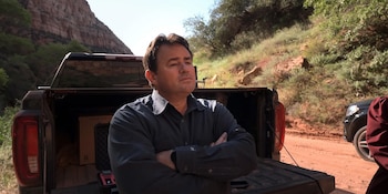 Hombre de mediana edad con camisa gris de manga larga y brazos cruzados, de pie frente a la caja abierta de una camioneta oscura, en un paisaje desértico rocoso