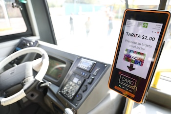 Interior de un autobús con volante y tablero de control; un validador de pasaje digital muestra "Tarifa $2.00" e instrucción de pasar tarjeta