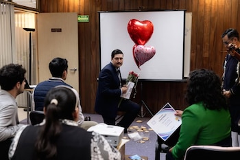 Hombre arrodillado en una oficina con un traje oscuro, sosteniendo rosas y una tarjeta, frente a globos de corazón. Hay personas observando y galletas en el suelo