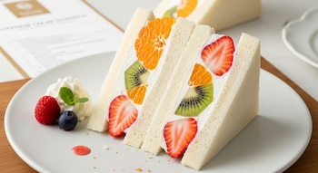 Sándwich de crema japonés triangular en plato blanco, con capas de pan, crema batida, fresas, kiwis y gajos de mandarina. Adornado con crema, frambuesa y arándano.