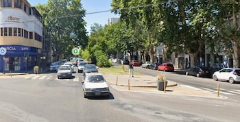 Calle urbana con vehículos circulando y estacionados, edificios con locales comerciales, arbolado y una persona en la acera