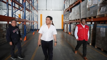Tres hombres caminan por el pasillo central de un almacén. Estantes industriales vacíos a la izquierda y estantes con mercadería paletizada a la derecha