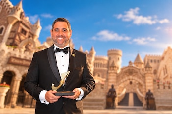 Un hombre con traje formal negro y corbata de lazo sostiene una estatuilla dorada frente a una edificación de estilo medieval