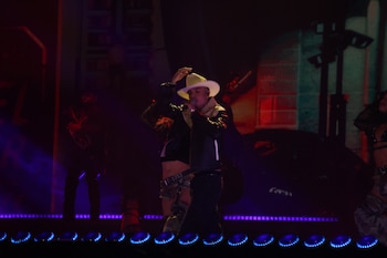 Un hombre con sombrero vaquero blanco canta en un micrófono sobre un escenario con luces y bailarines al fondo
