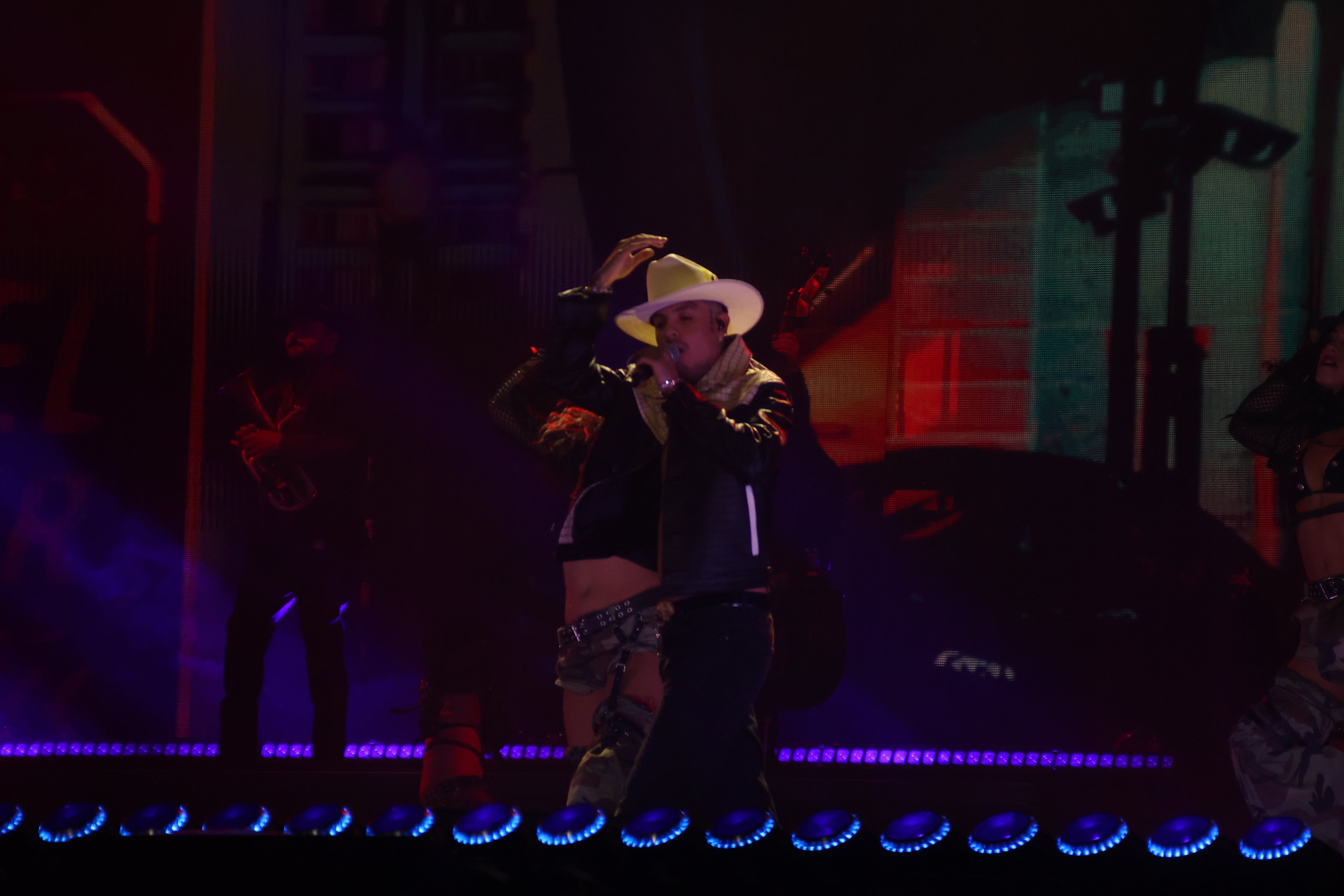 Un cantante con sombrero blanco y chaqueta oscura interpreta una canción durante una presentación en un escenario iluminado. (Selene Miranda/Infobae México)