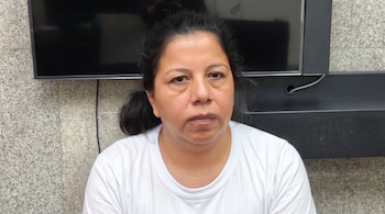 Mujer con cabello oscuro y camiseta blanca sentada frente a una pared de piedra clara, con un televisor apagado y una barra de sonido detrás.