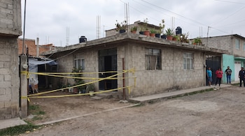 Casa de concreto con cinta amarilla en el frente, plantas en la azotea y cuatro personas de pie en una calle no pavimentada.