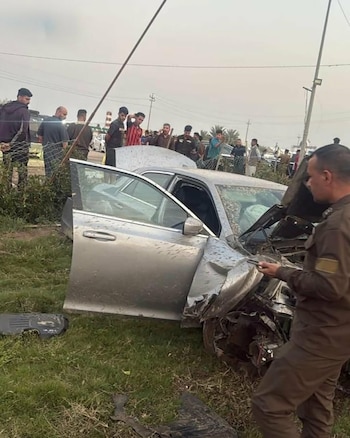 Coche plateado dañado con el capó abierto y airbags desplegados en un campo cubierto de hierba. Varios hombres se agrupan alrededor del vehículo