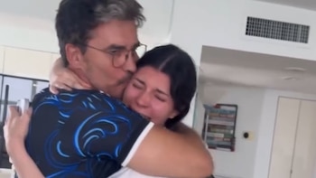 Un hombre con gafas y camisa oscura abraza a una mujer de cabello oscuro que llora. El hombre besa la mejilla de la mujer. Fondo de interior claro