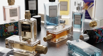 Múltiples frascos de perfume genéricos, transparentes y de diversas formas, con líquidos ámbar, azules y rosas, dispersos sobre una superficie, algunos abiertos.