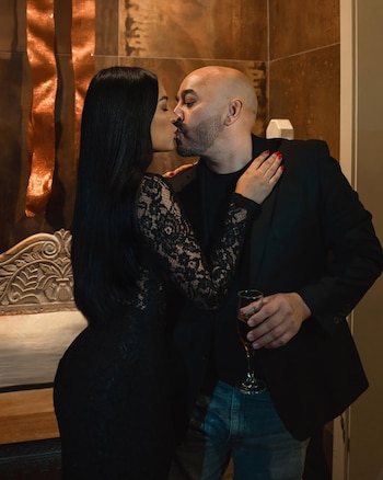 Lupillo Rivera y Taína Pimentel se besan apasionadamente. Él, calvo, con barba y en saco negro, sostiene una copa. Ella, con pelo largo oscuro, viste un vestido de encaje negro
