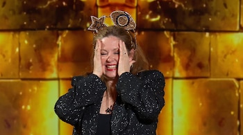 Mujer de mediana edad con cabello castaño, diadema dorada con estrella y ojo, chaqueta negra de lentejuelas, manos cubriendo su cara, y boca abierta sonriendo.